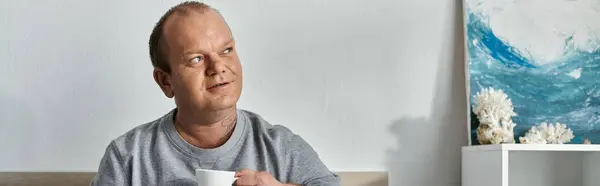 Человек с инклюзивностью сидит в комнате, держа чашку, и смотрит вверх, затерянный в мыслях, с картиной, изображающей волны на заднем плане. — стоковое фото