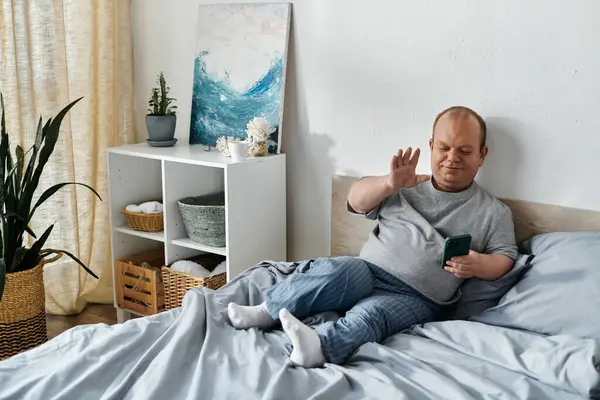 A man with inclusivity sits on a bed waving at a phone. — Stock Photo