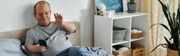 Un uomo con inclusività siede sul suo letto, sorridendo e salutando mentre tiene in mano uno smartphone. — Foto stock