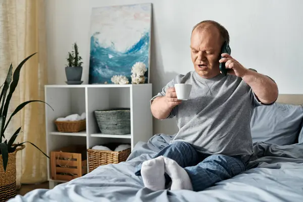 A man with inclusivity sits on his bed, holding a cup of coffee and talking on the phone. — Stock Photo