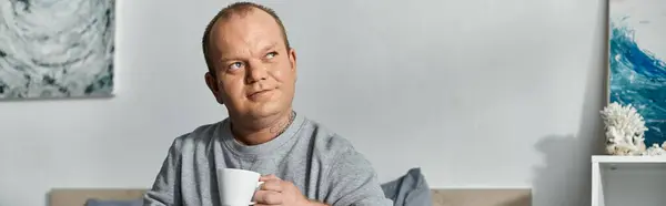 Um homem com inclusividade em uma camisola cinza senta-se em um sofá, segurando uma caneca de café. — Fotografia de Stock
