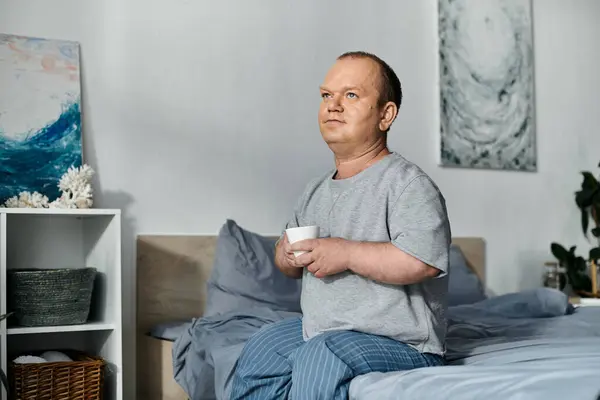 A man with inclusivity sits on his bed in the morning, holding a cup and looking thoughtfully out of frame. — Stock Photo