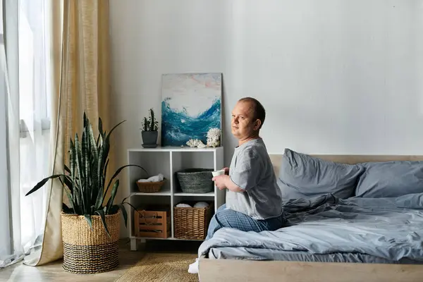 A man with inclusivity sits on his bed, holding a cup, in a bright room with a plant and a painting. — Stock Photo
