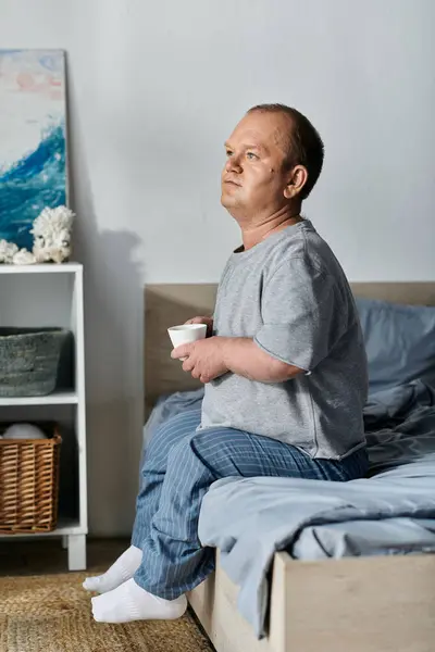 Un hombre con inclusividad se sienta en el borde de una cama, sosteniendo una taza, y mirando por la ventana. - foto de stock