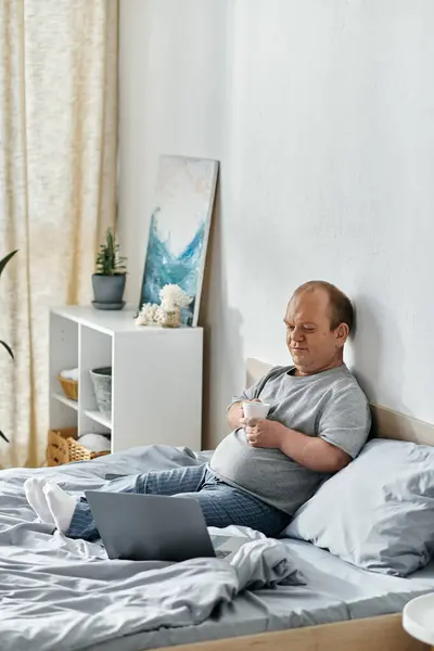 A man with inclusivity sits in bed, holding a cup of coffee, and using a laptop. — Stock Photo