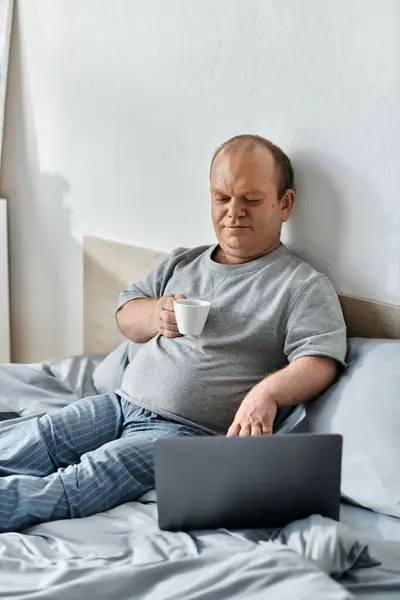 Un uomo con inclusività si siede a letto, gustando una tazza di caffè mentre lavora sul suo computer portatile. — Foto stock