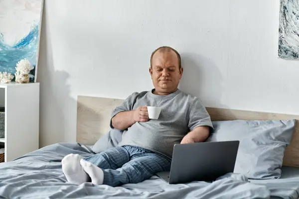A man with inclusivity sits in bed, enjoying a cup of coffee while using his laptop. — Stock Photo