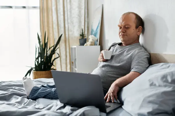 A man with inclusivity in pajamas sits in bed with a laptop and a cup of coffee. — Stock Photo