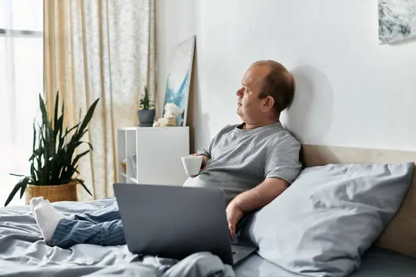 A man with inclusivity sits in bed, holding a cup and looking out the window, with a laptop resting on his lap. — Stock Photo