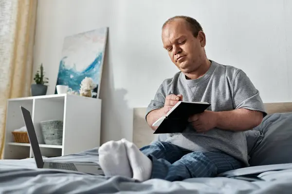 A man with inclusivity sits on his bed, writing in a notebook, with a laptop beside him. — Stock Photo