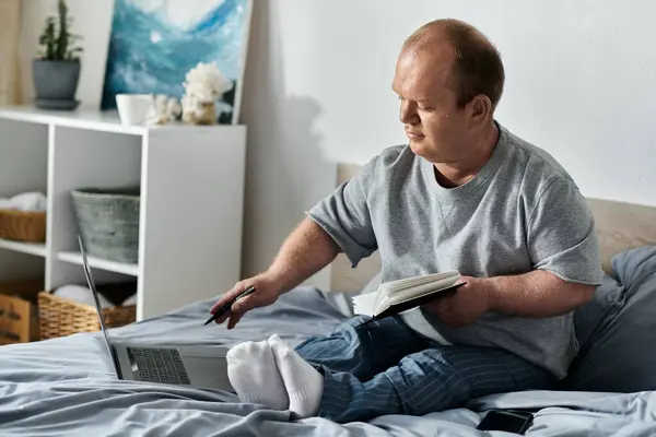 Ein inklusiver Mann sitzt auf seinem Bett, arbeitet an einem Laptop und liest ein Buch. — Stockfoto