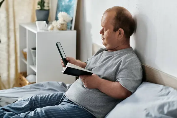 Ein inklusiver Mann sitzt im Bett und schaut auf sein Handy, während er ein Buch in der Hand hält. — Stockfoto