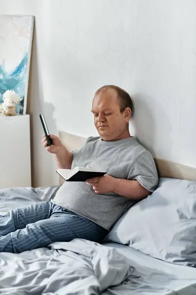 Человек с инклюзивностью сидит в постели, читает книгу и держит телефон.. — стоковое фото