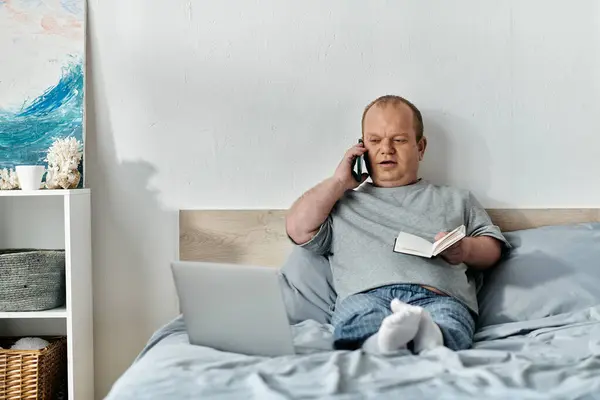A man with inclusivity sits in bed, talking on the phone and making notes in a notebook. — Stock Photo