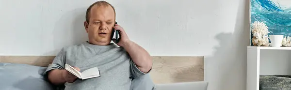 A man with inclusivity sits on a bed, talking on the phone and reading a book. — Stock Photo