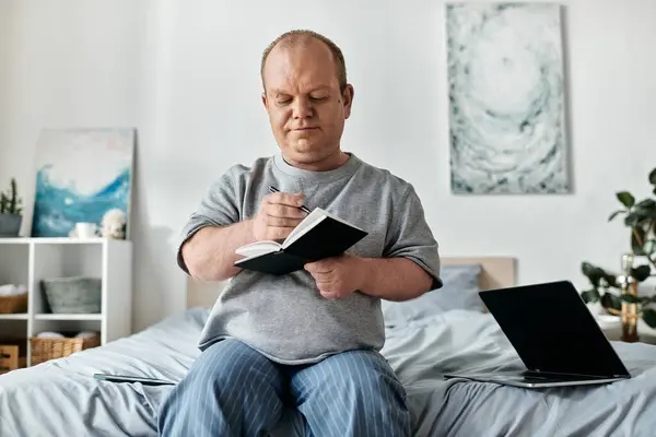 Un homme avec inclusivité est assis sur un lit écrit dans un cahier, avec un ordinateur portable à proximité. — Photo de stock