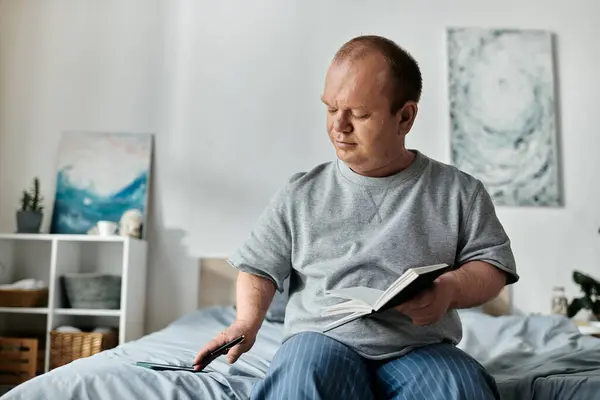 A man with inclusivity sits on the edge of his bed, reading a book and looking at his phone. — Stock Photo