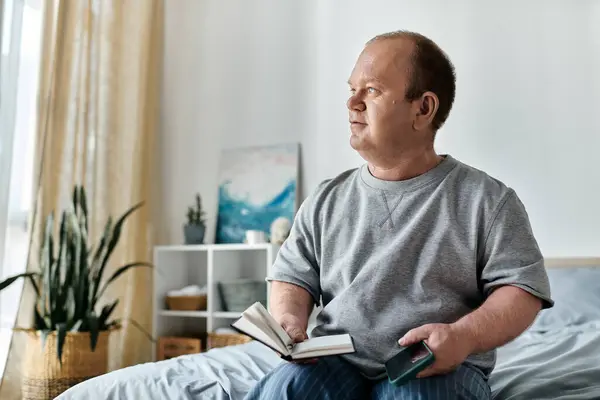 Ein inklusiver Mann sitzt auf einem Bett, blickt aus dem Fenster und hält ein Notizbuch und ein Telefon in der Hand.. — Stockfoto