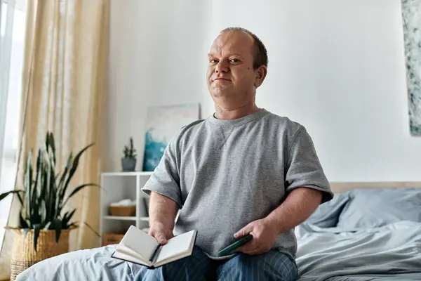 Un hombre con inclusividad se sienta en una cama, sosteniendo un libro y un teléfono, mirando pensativamente hacia adelante. - foto de stock