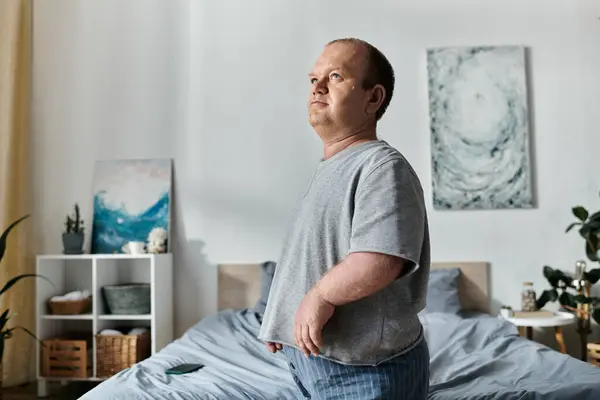 Человек с инклюзивностью стоит у кровати, задумчиво глядя в окно. — стоковое фото