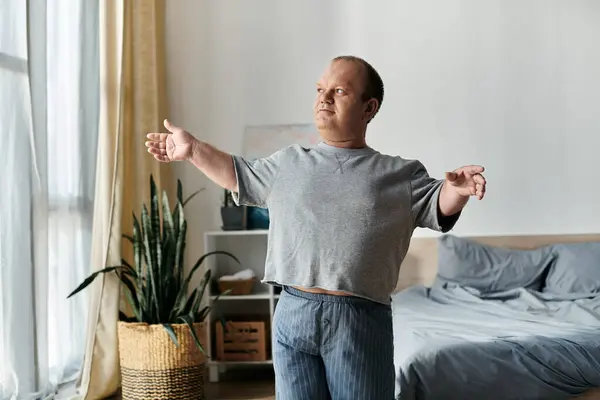 A man with inclusivity stretches his arms out in his pajamas while standing by a window. — Stock Photo