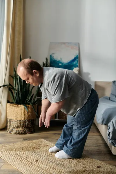 A man with inclusivity in a grey shirt and blue pants stretches on a mat in his living room. — Stock Photo