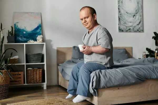 Um homem com inclusão senta-se em uma cama, segurando uma xícara de café e olhando para a câmera. — Fotografia de Stock