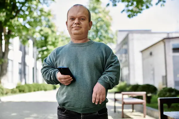 A man with inclusivity holding a phone walks along a city street with green trees in the background. — Stock Photo