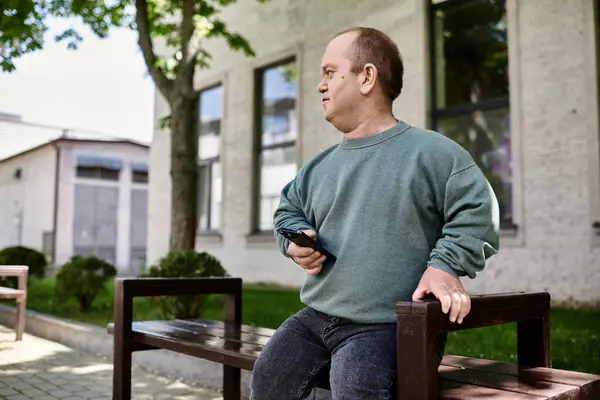 A man with inclusivity sits on a bench, holding a phone, in front of a building. — Stock Photo