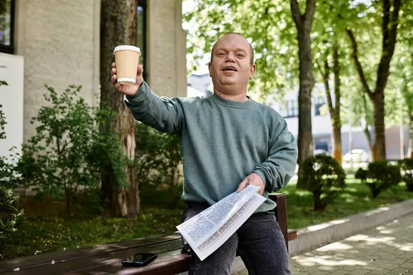 A man with inclusivity sits on a park bench, holding a cup of coffee and a newspaper, with a smile on his face. — Stock Photo