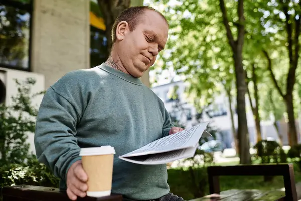 Человек с инклюзивностью сидит на скамейке, читает газету и наслаждается чашкой кофе.. — стоковое фото