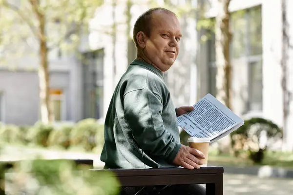 Un hombre con inclusividad se sienta en un banco, lee un periódico y disfruta de una taza de café. - foto de stock