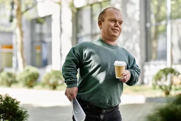 Un homme inclusif marche dans une rue de la ville, portant une tasse de café et un document plié. — Photo de stock