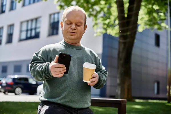 A man with inclusivity sits on a bench, using his phone and holding a takeaway coffee cup. — Stock Photo