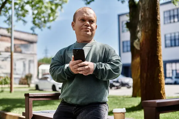 A man with inclusivity sits on a bench in a city park, using his smartphone while enjoying a cup of coffee. — Stock Photo