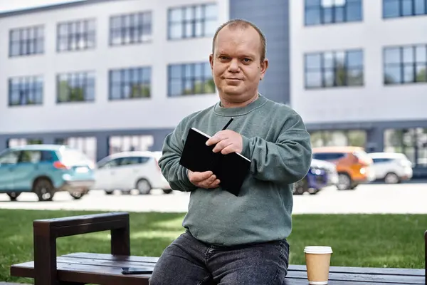 A man with inclusivity sits on a bench outside a building, holding a notebook and pen. He appears relaxed and content, taking notes on his day. — Stock Photo