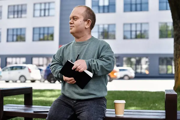 A man with inclusivity sits on a bench in a city park, thoughtfully writing in a notebook with a coffee beside him. — Stock Photo