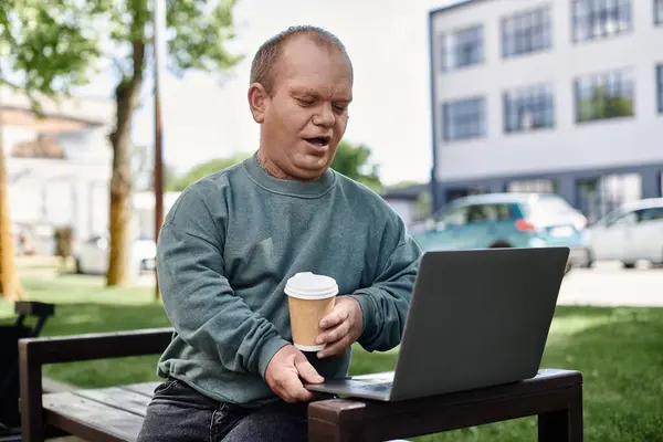 Un uomo con inclusività con inclusività siede su una panchina in un parco, lavora sul suo computer portatile e si gode un caffè. — Foto stock