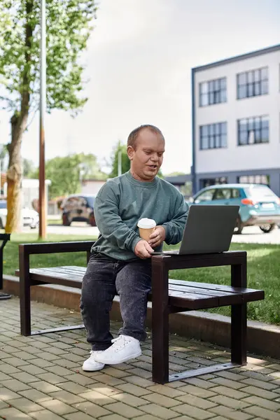 A man with inclusivity sits on a bench in a park, enjoying a cup of coffee and working on his laptop. — Stock Photo
