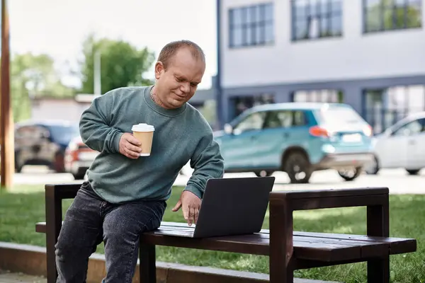 Un homme avec inclusivité est assis sur un banc de parc, dégustant du café et utilisant un ordinateur portable par une journée ensoleillée. — Photo de stock