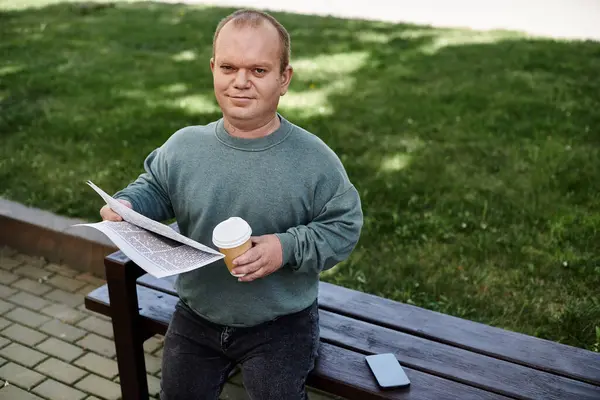 Un hombre con inclusividad se sienta en un banco del parque, leyendo un periódico y sosteniendo una taza de café. - foto de stock