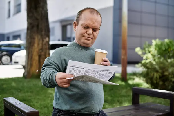 Человек с инклюзивностью сидит на скамейке, наслаждаясь утренним кофе и читая газету. — стоковое фото