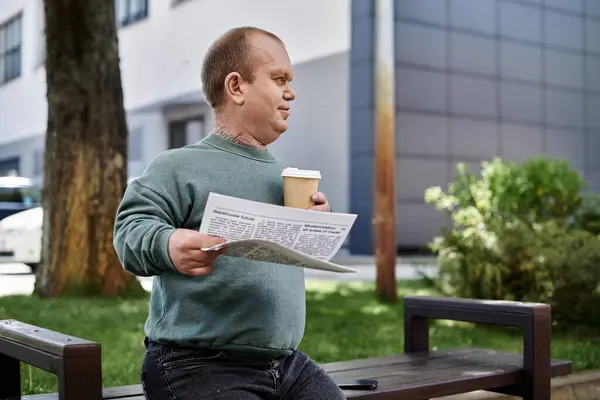 Человек с инклюзивностью сидит на скамейке в парке, читает газету и держит чашку кофе.. — стоковое фото