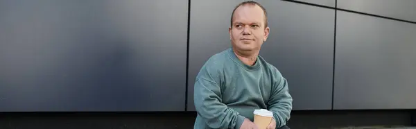 A man with inclusivity sits casually in front of a modern building, holding a cup of coffee. — Stock Photo