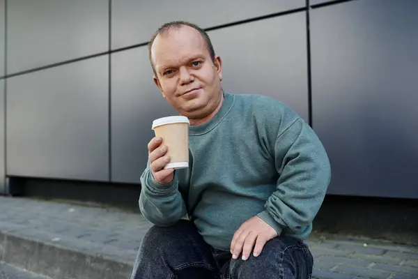 A man with inclusivity sits on a curb, holding a coffee cup and smiling for the camera. — Stock Photo