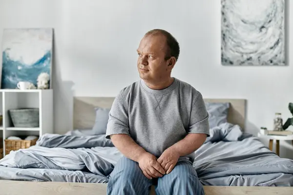 A man with inclusivity sits on the edge of a bed in a bedroom, looking thoughtful. — Stock Photo