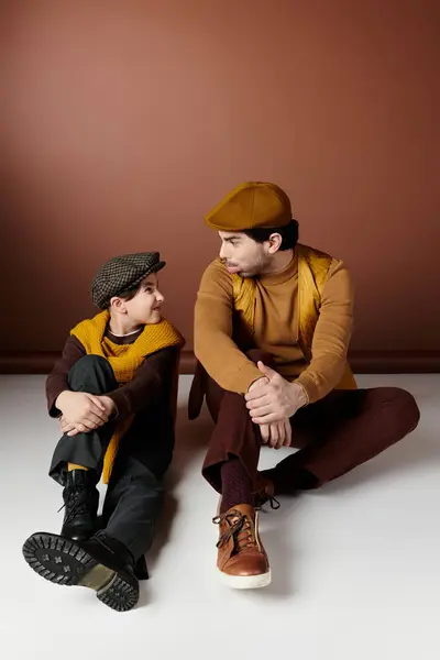 A father and son sit on a white floor, dressed in brown and yellow clothing, smiling at each other. — Stock Photo