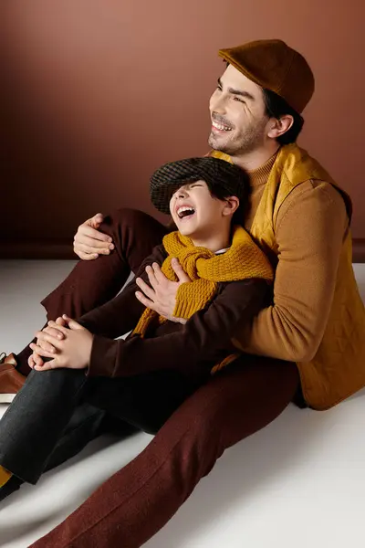 Um pai e um filho sentam-se em um chão branco na frente de uma parede marrom, rindo juntos. — Fotografia de Stock