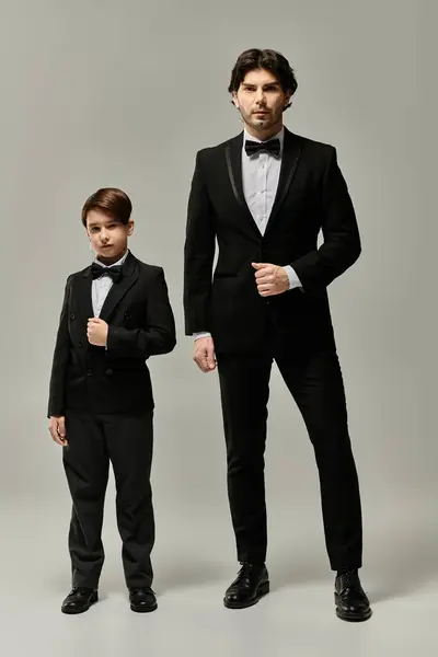A father and his son, both wearing black tuxedos, stand side by side against a gray background. — Stock Photo