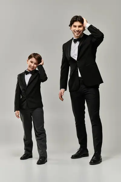 A father and son, both wearing black suits and bowties, stand side-by-side against a gray backdrop. — Stock Photo